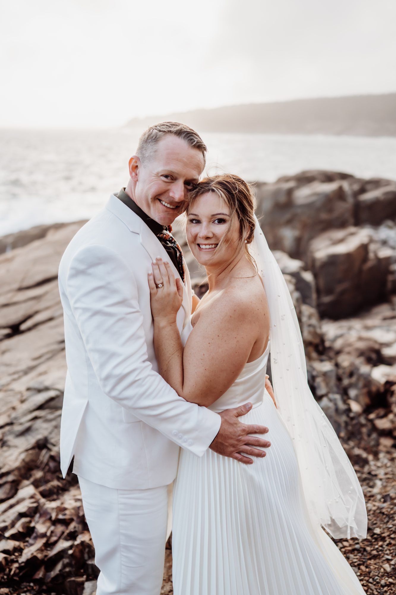 Bride and groom embracing along rocky shoreline at Thunder Hole in Acadia.
