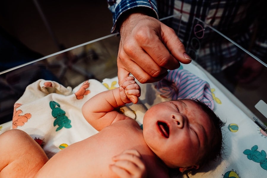 Newborn baby laying in warmer holding fathers finger