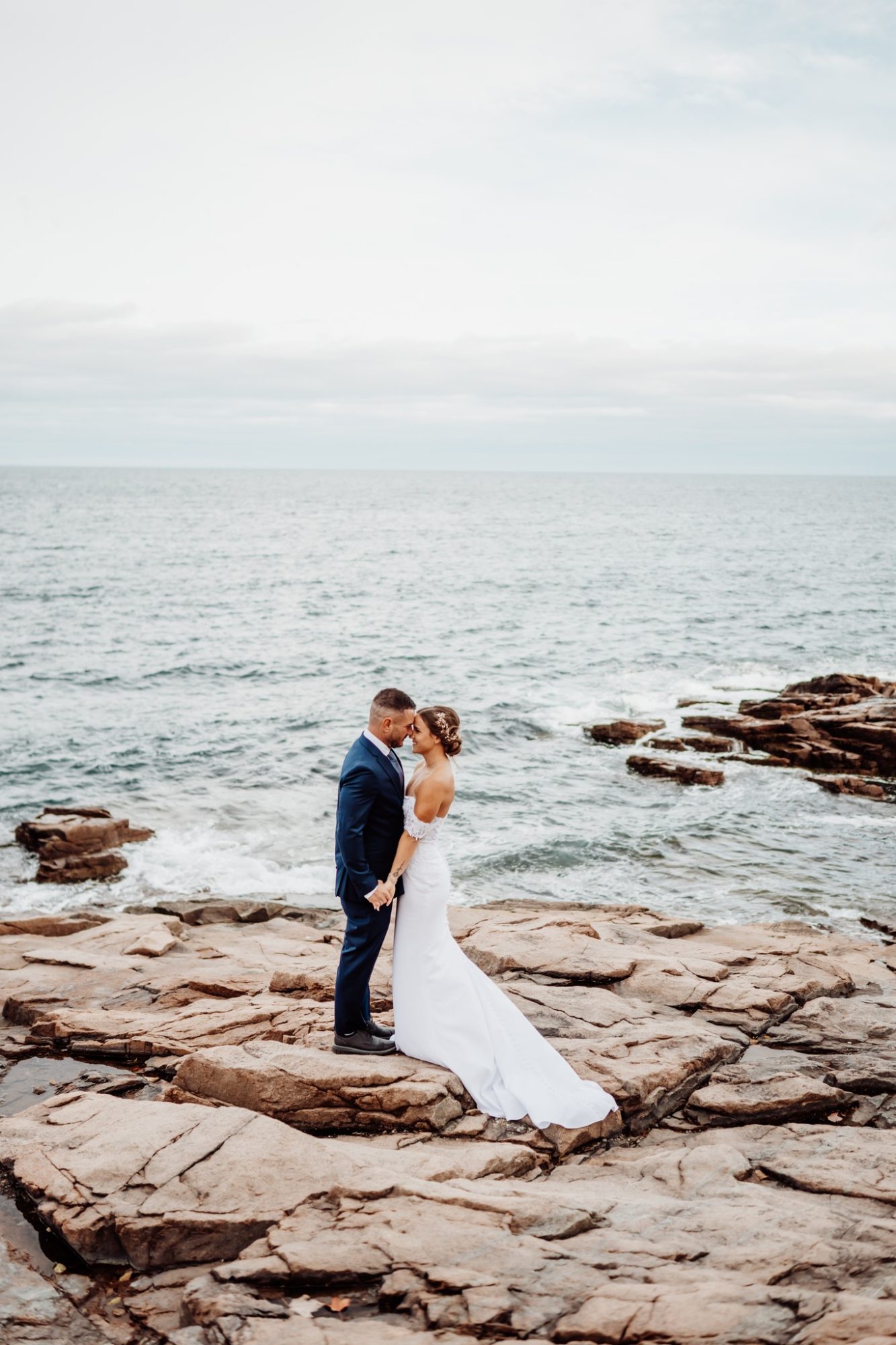 Newly married couple standing on the rocky shoreline at Schooner Head Overlook in Acadia National Park, waves crashing against the Maine coast during their intimate elopement.