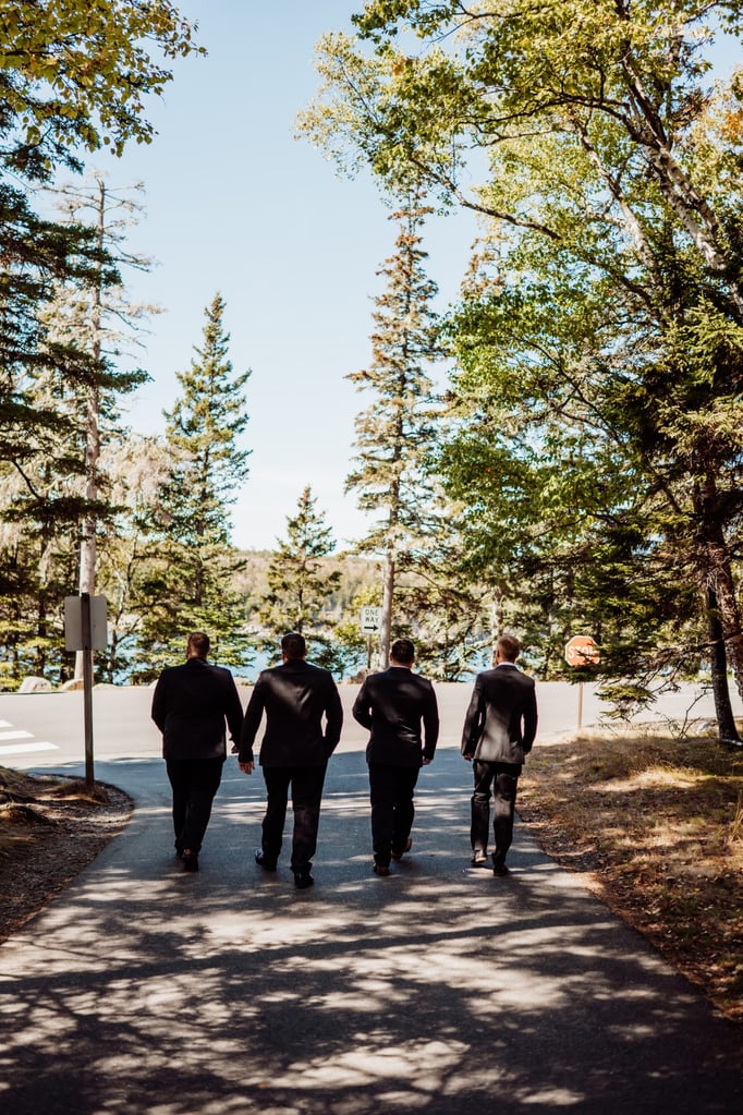 Four groomsmen walking down a tree-lined path toward the water at Otter Point in Acadia National Park, heading to the wedding ceremony on a sunny fall afternoon.