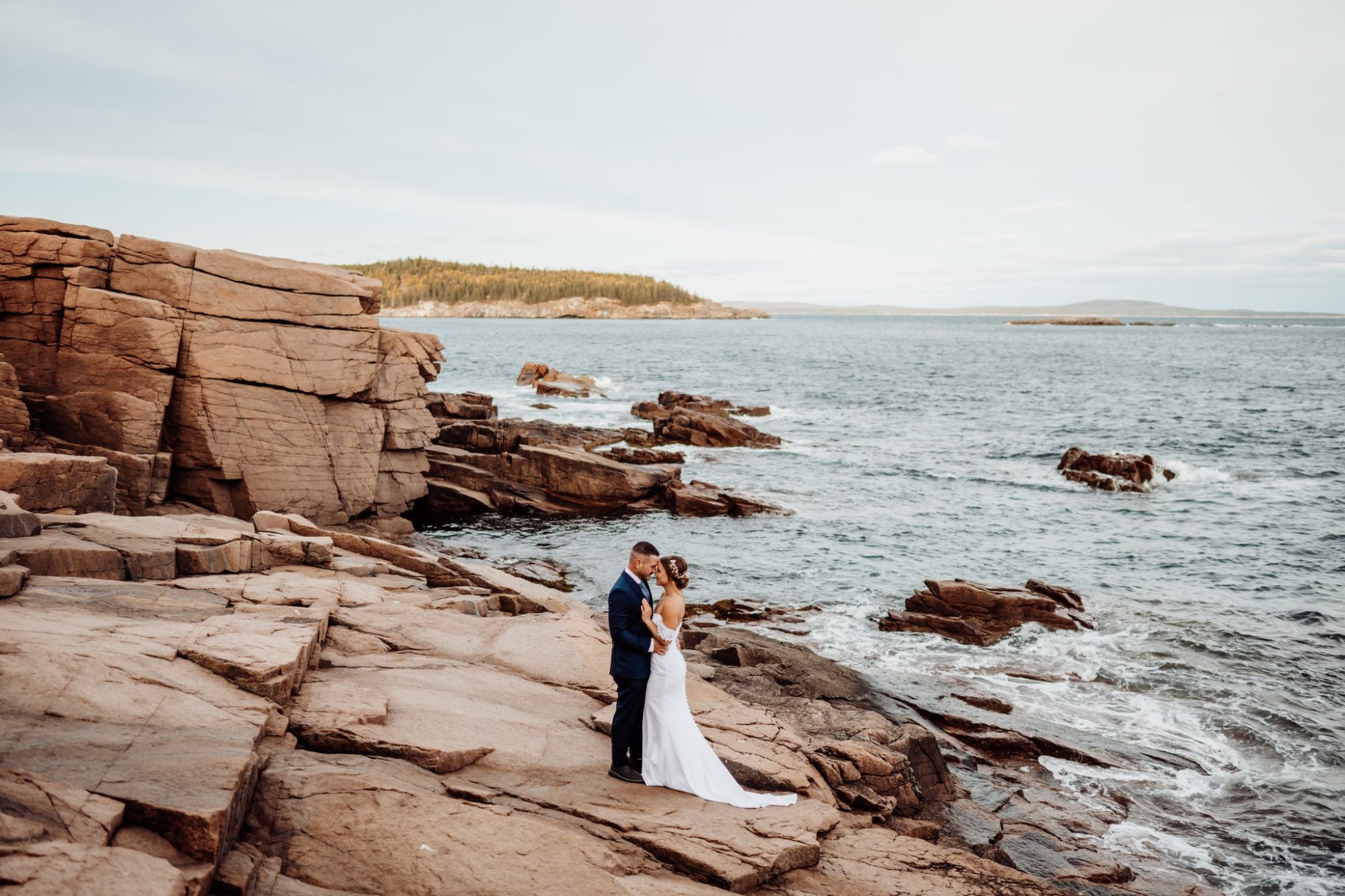 Bride and groom embracing on granite cliffs at Schooner Head Overlook in Acadia National Park above the Atlantic Ocean.