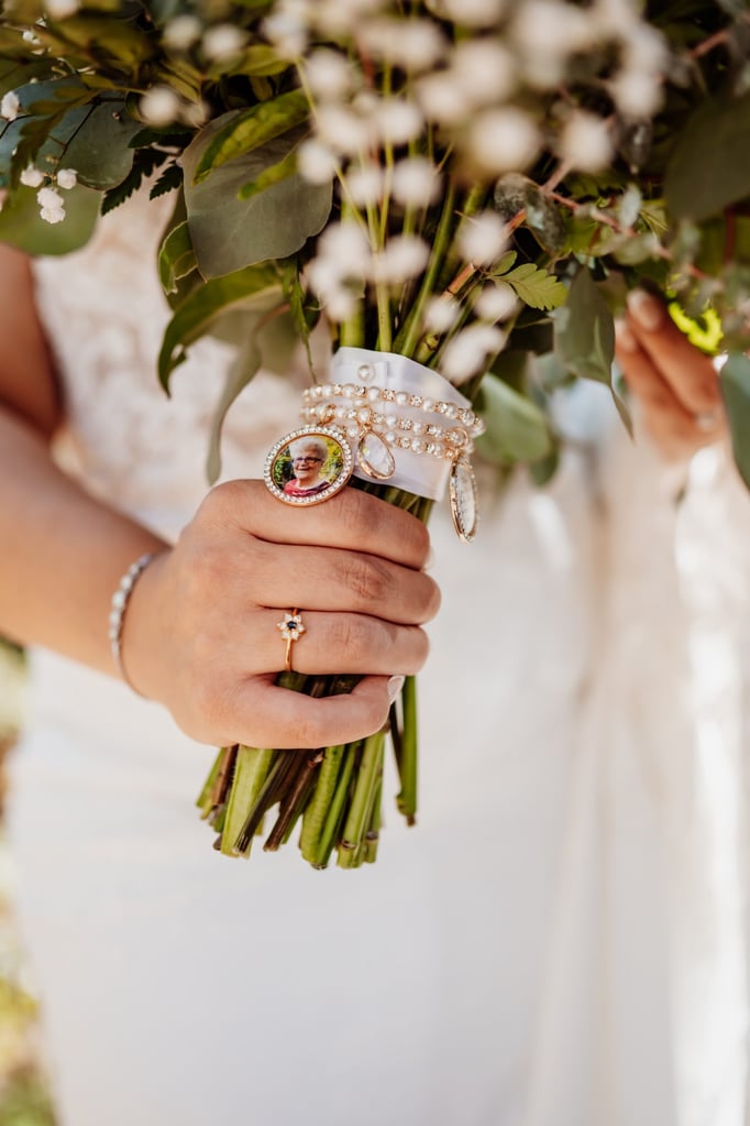 Close-up of a bride holding her bouquet wrapped with pearl and rhinestone charms featuring a photo of a loved one, captured during an Acadia National Park wedding in Bar Harbor, Maine.