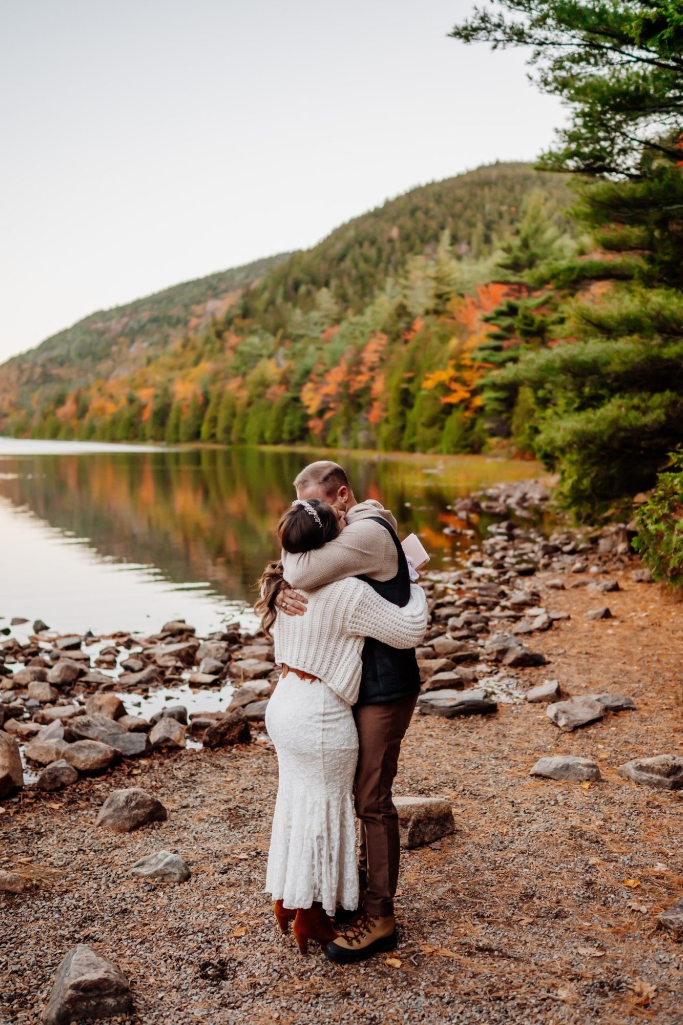Bride and groom hugging beside Jordan Pond with colorful fall mountains.