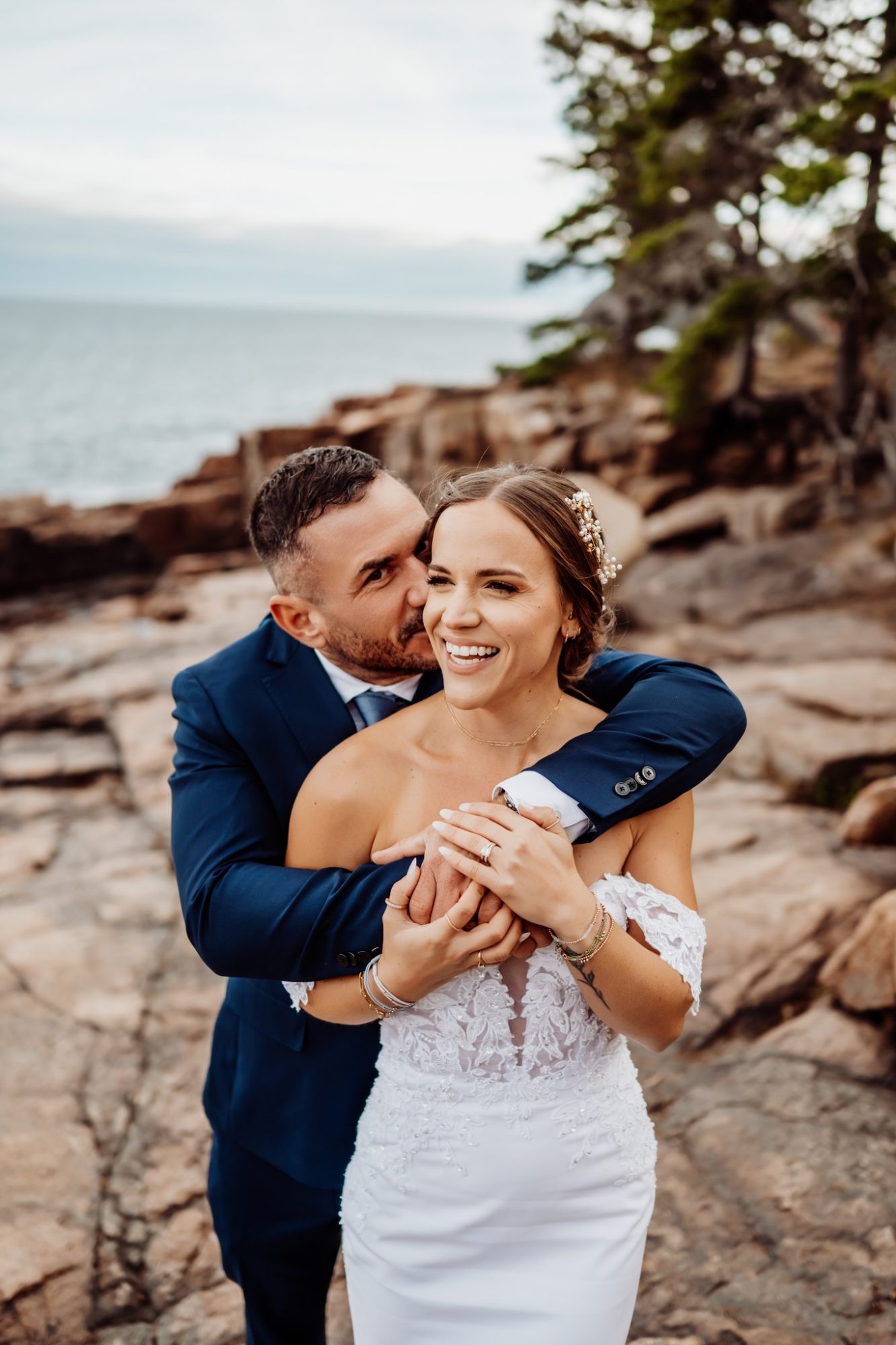 Groom hugging bride from behind as she laughs on the rocky shoreline during their Schooner Head Overlook elopement in Acadia National Park.