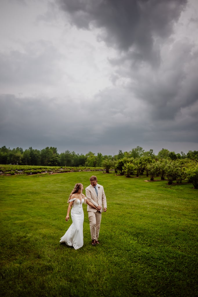 Bride and groom walking across a grassy field with dramatic storm clouds rolling in overhead, creating a moody and romantic atmosphere.