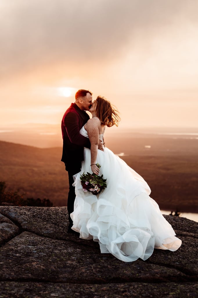 Bride and groom sharing a romantic kiss on Cadillac mountain summit at sunset during their elopement, flowing dress and warm golden light