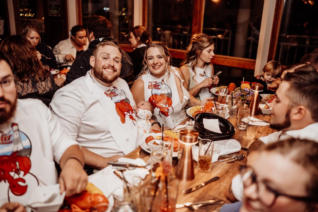 Bride and groom wearing lobster bibs and smiling while enjoying a traditional Maine lobster dinner with guests after their Acadia National Park wedding.