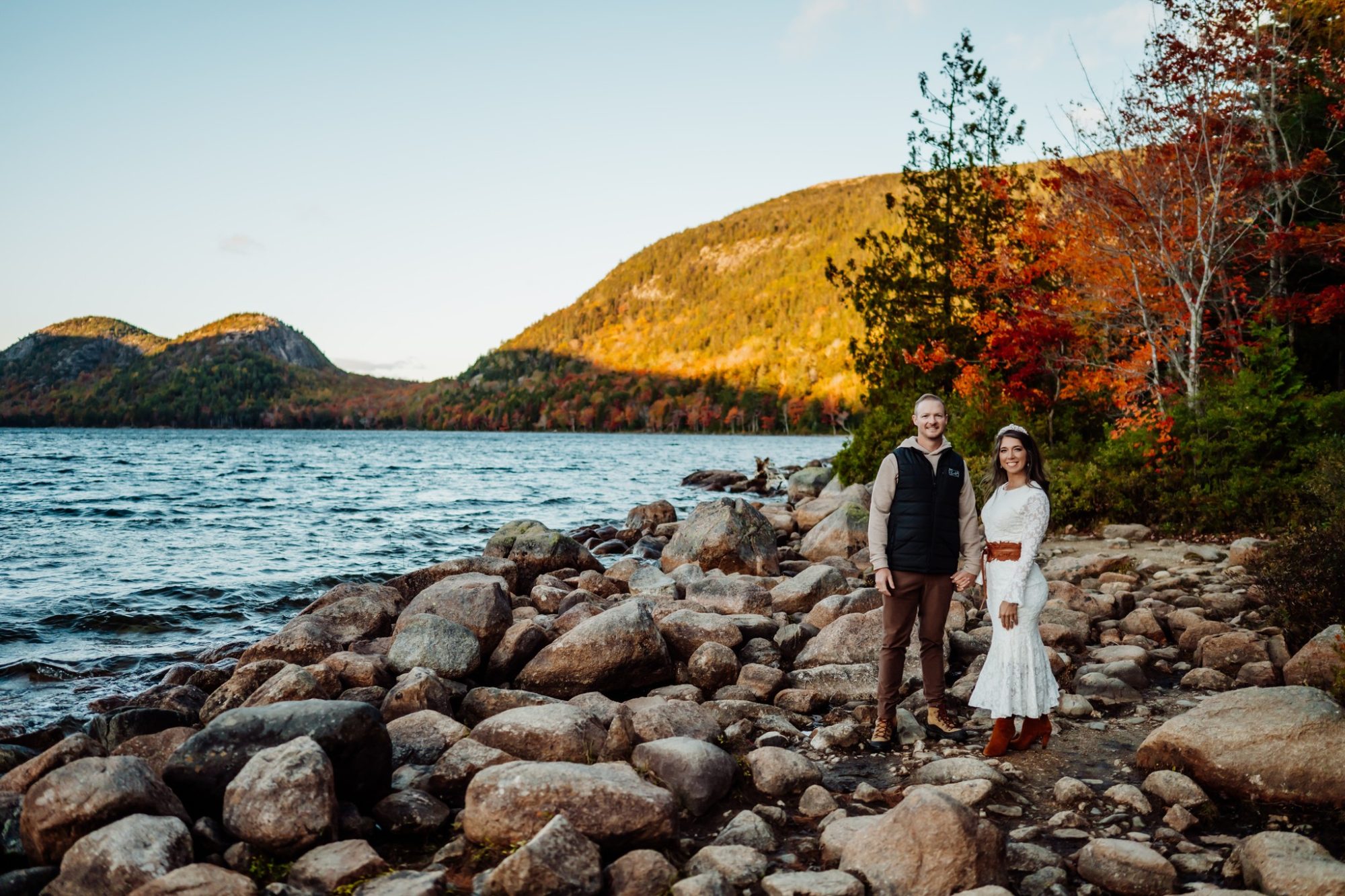 Couple on rocky shoreline with colorful fall mountain backdrop in Acadia.