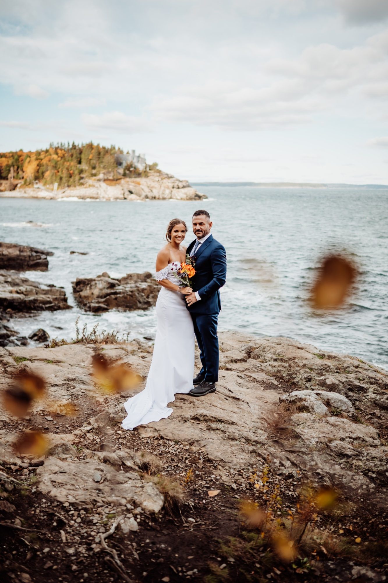 Bride and groom posing with bouquet of orange roses at Schooner Head Overlook in Acadia National Park overlooking the Atlantic Ocean.
