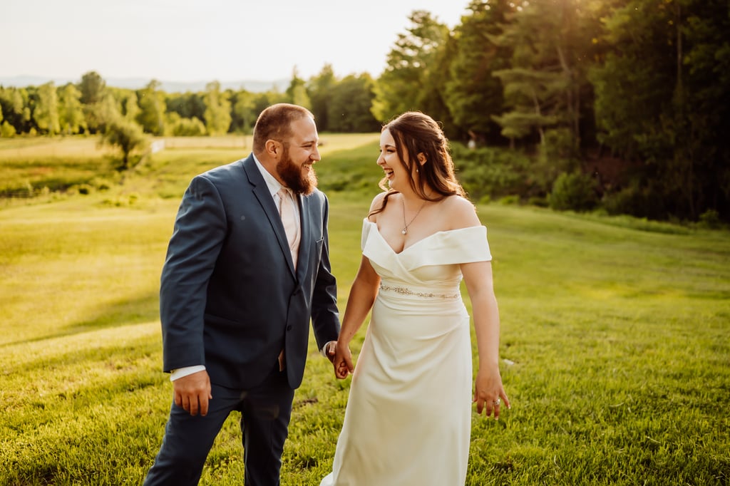 The bride and groom walk hand in hand in a field at sunset.