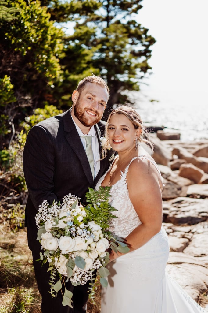 Bride and groom smiling together after their Acadia National Park wedding, holding a lush white and green bouquet with the ocean behind them.