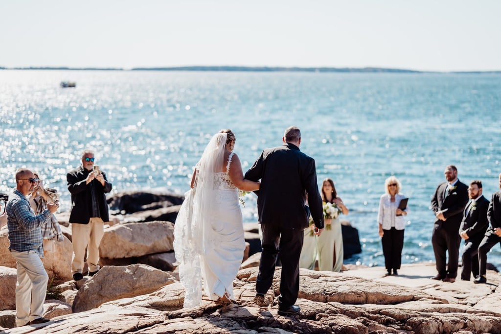 Bride walking arm-in-arm with her father across the sunlit cliffs of Otter Point in Acadia National Park, guests capturing the moment against the sparkling ocean backdrop.
