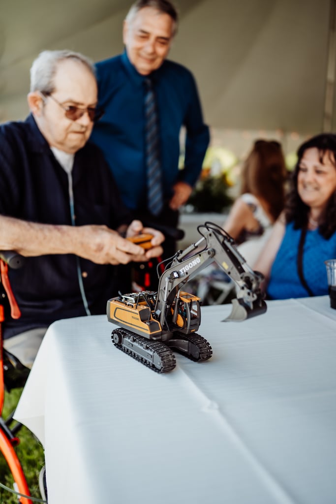 Two older men, one operating a remote-controlled toy excavator, sharing a moment of fun at a wedding reception.