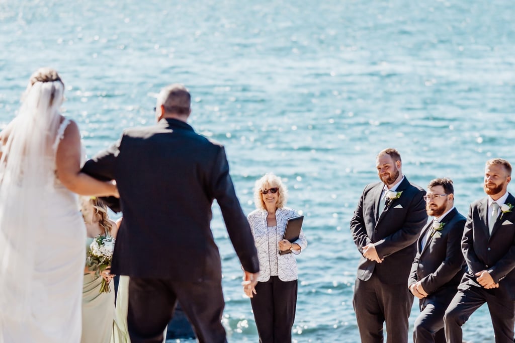 Bride and her father walking toward the groom and officiant on the rocky shore of Acadia National Park, surrounded by family and friends overlooking the Atlantic Ocean.