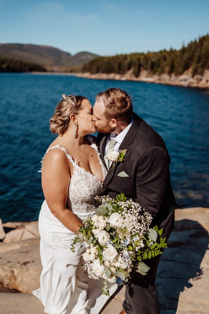 Bride and groom sharing a kiss in front of the ocean at Otter Point in Acadia National Park, holding a bouquet of white roses and greenery under clear blue skies.