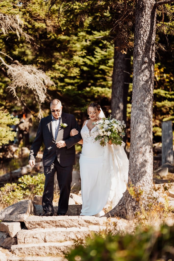 Bride in an elegant lace gown walking arm-in-arm with her father down stone steps through the forest toward her Acadia National Park wedding ceremony, bouquet of white flowers in hand.