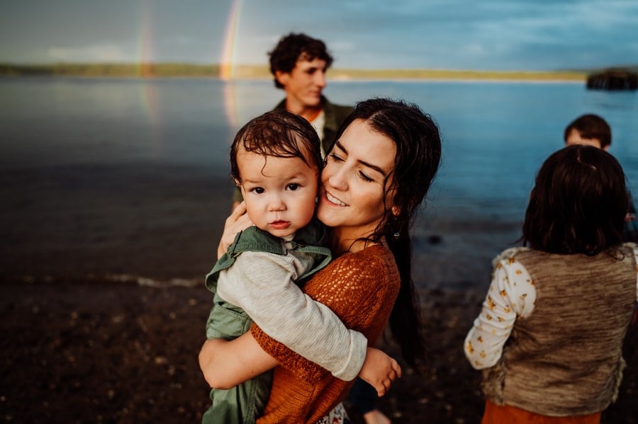Mother in orange knit top holding baby boy wearing olive green overalls in front of water and double rainbow