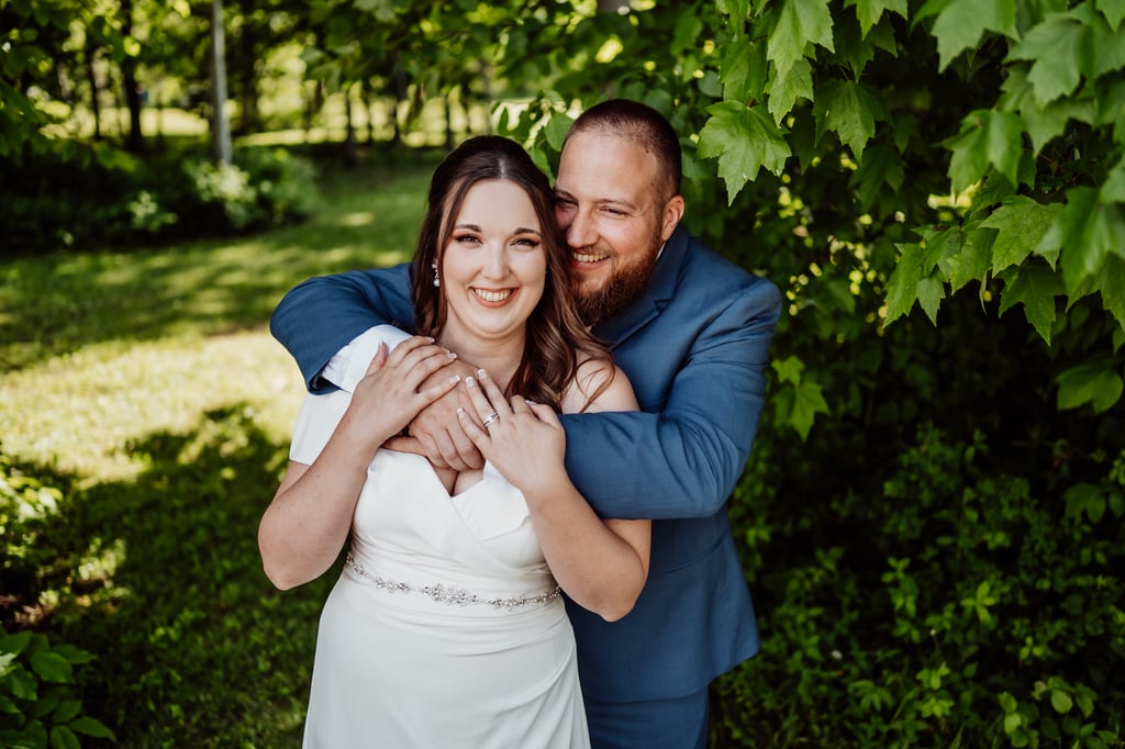 A bride and groom embracing and smiling happily, standing outdoors with green foliage in the background.