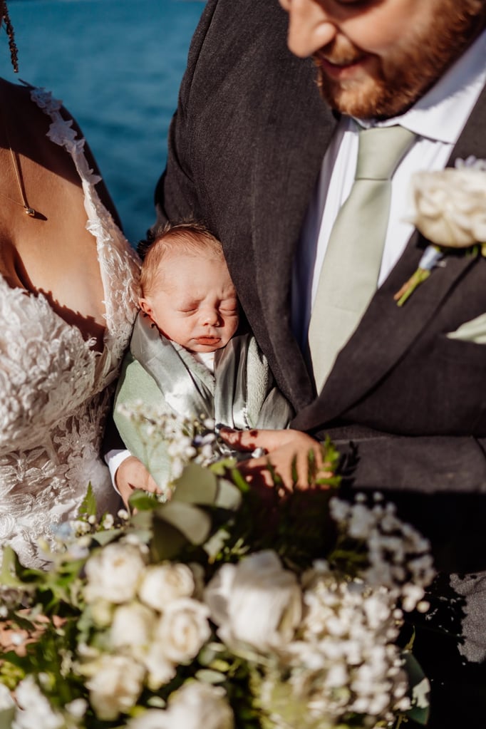 Bride and groom holding their newborn baby wrapped in soft sage fabric, surrounded by white flowers and the coastal backdrop of Acadia National Park.