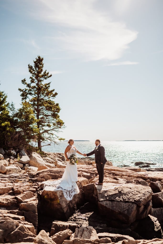 Bride and groom holding hands across the dramatic pink granite cliffs of Acadia National Park, ocean waves glimmering in the background during their Maine coast elopement.