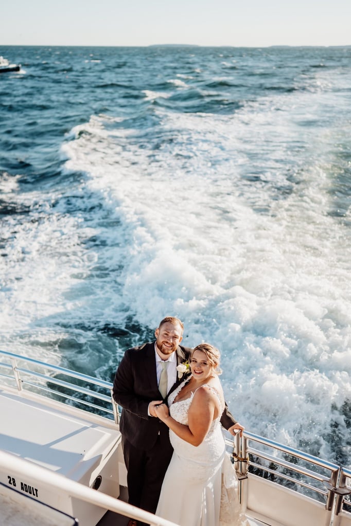 Bride and groom holding hands at the back of the boat, smiling as ocean waves trail behind them during their Bar Harbor sunset cruise wedding.