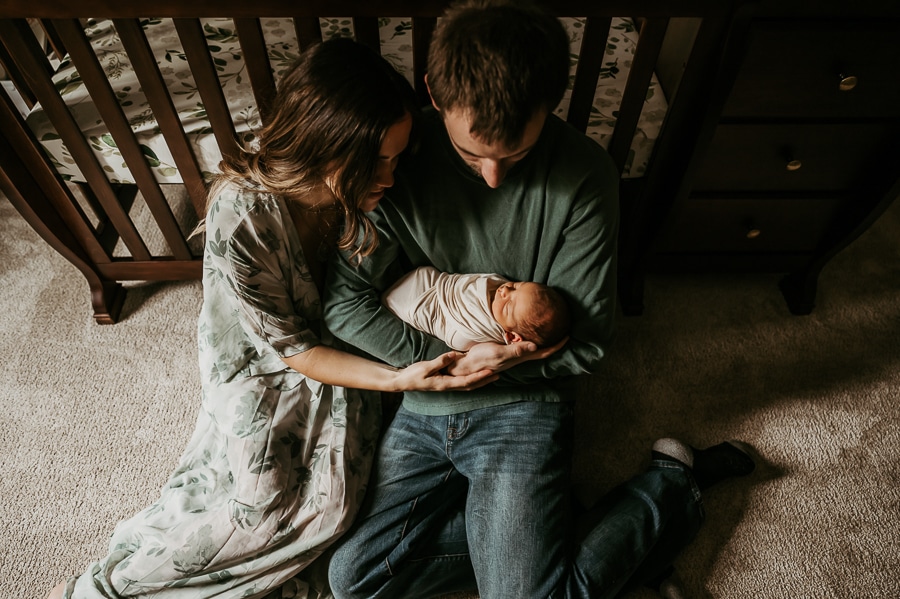 Mother and father sitting on floor with newborn in front of crib