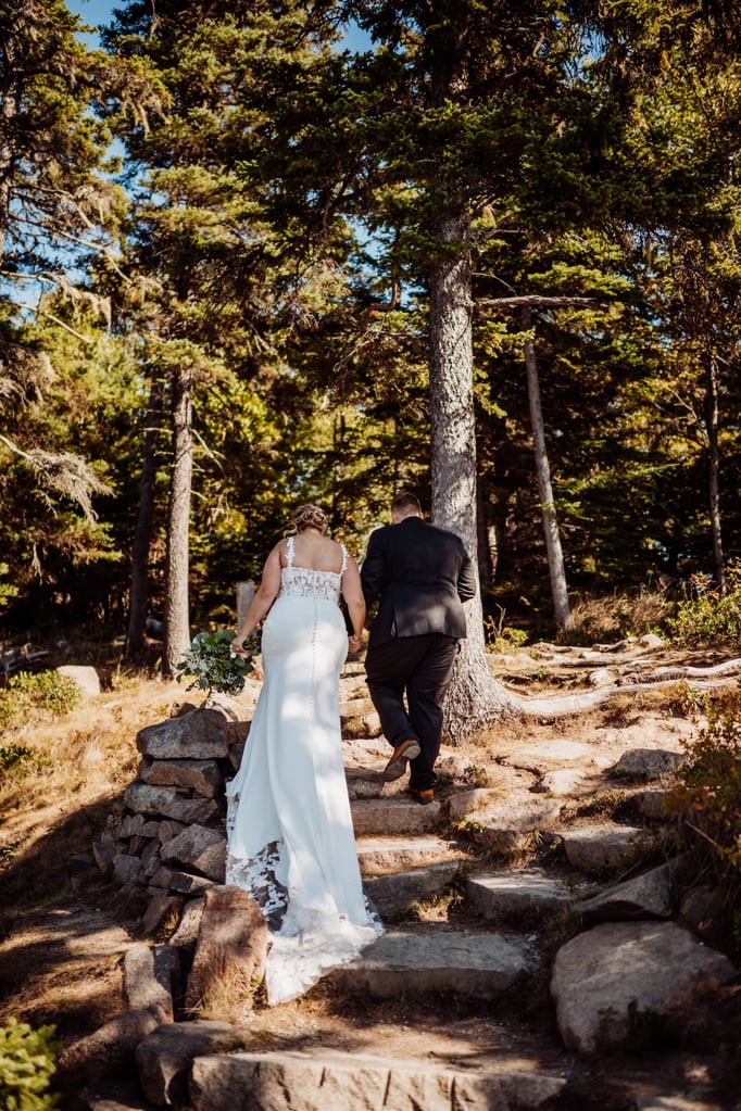Bride and groom walking hand-in-hand up a forest trail in Acadia National Park after their cliffside wedding ceremony.