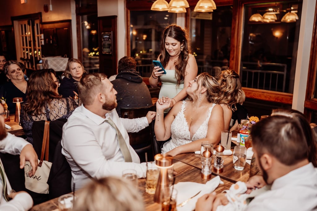 Bridesmaid giving a heartfelt toast while the bride and groom smile and hold hands at their waterfront restaurant reception in Bar Harbor.