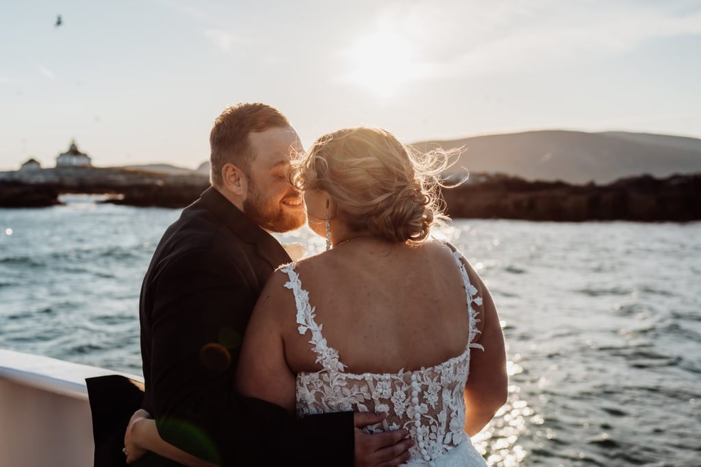 Bride and groom embracing at sunset on their Bar Harbor wedding cruise, with the lighthouse and rocky coastline in the distance.
