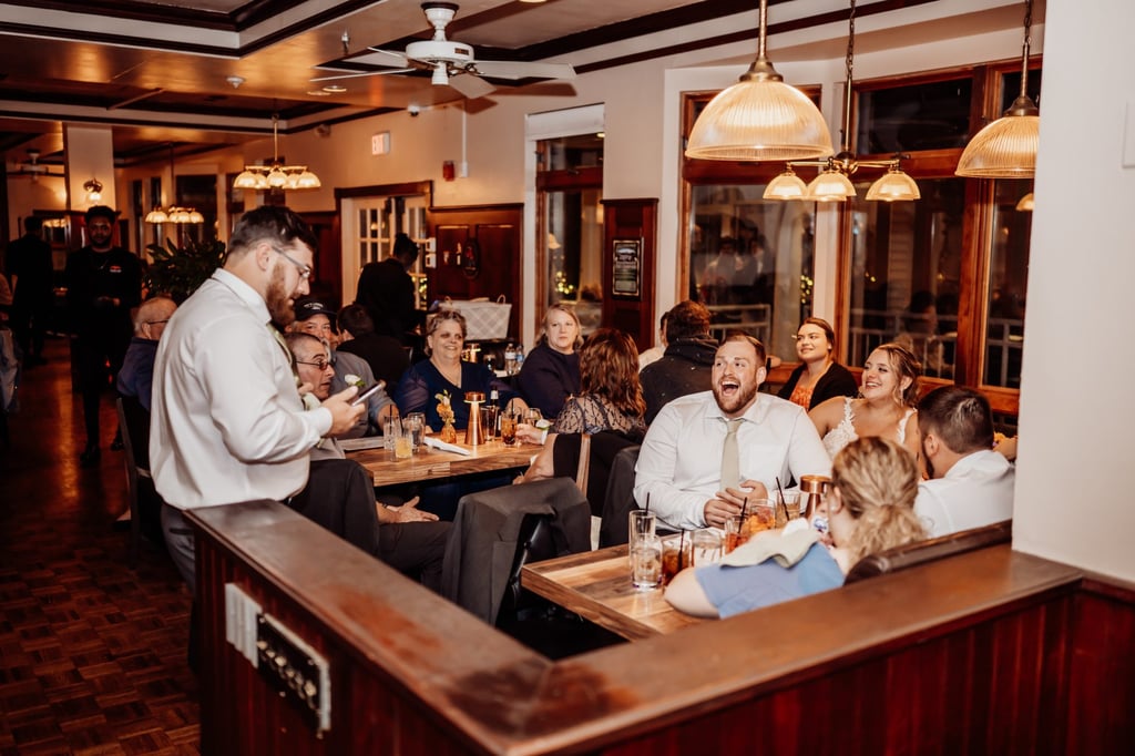 Best man giving a lively toast while the groom laughs and guests look on inside a cozy Maine coastal restaurant.