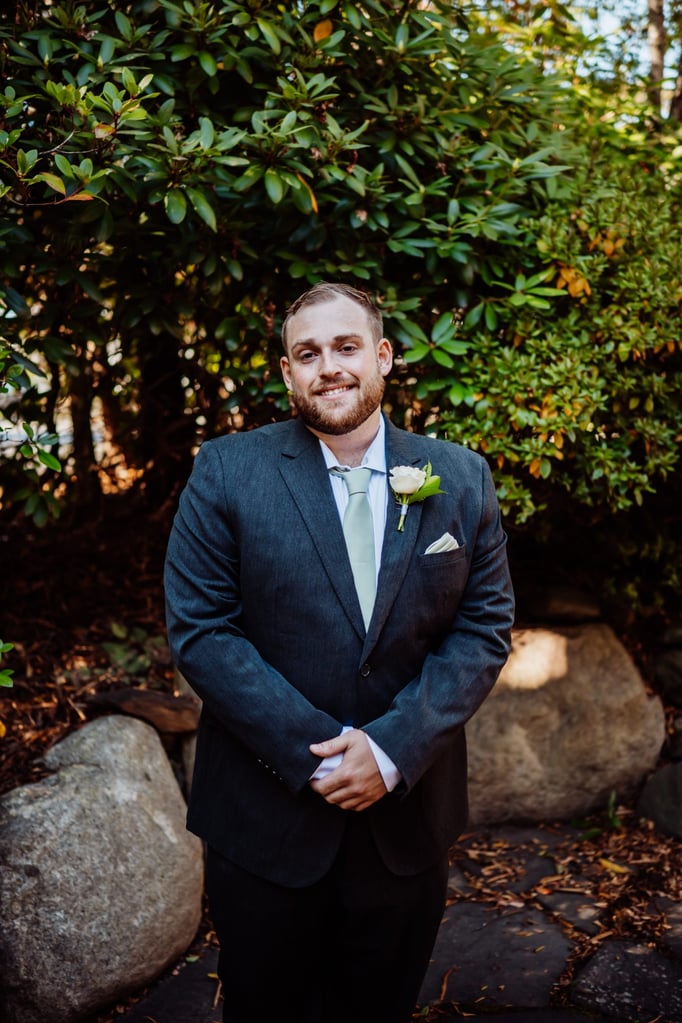 Groom standing confidently outdoors in a charcoal suit and light green tie, surrounded by greenery and natural stone.