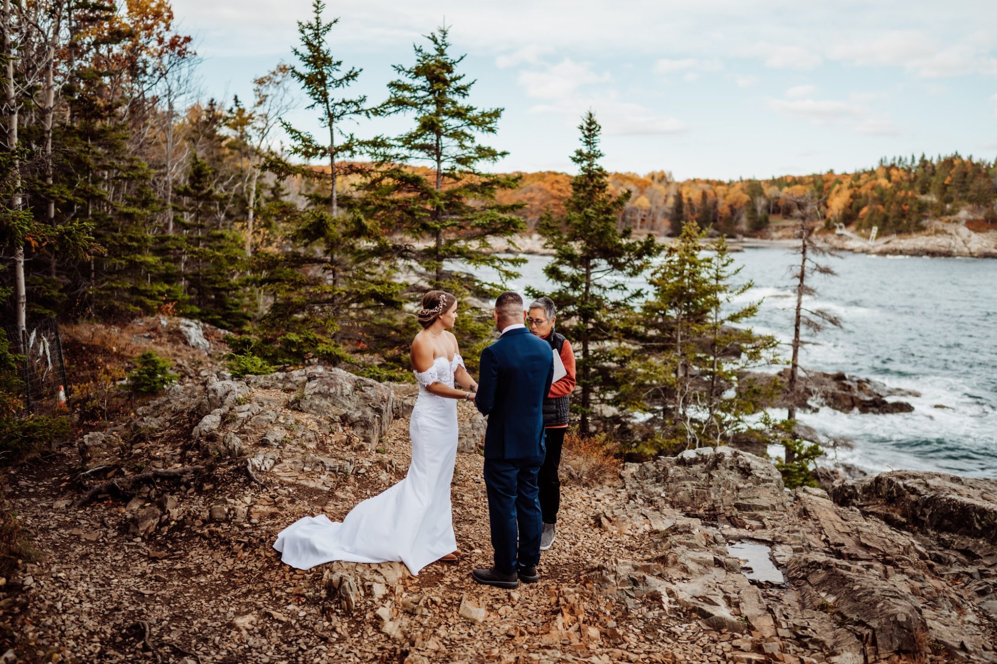 Bride and groom exchanging vows during a private elopement ceremony at Schooner Head Overlook in Acadia National Park, surrounded by rocky coastline and evergreen trees in fall.