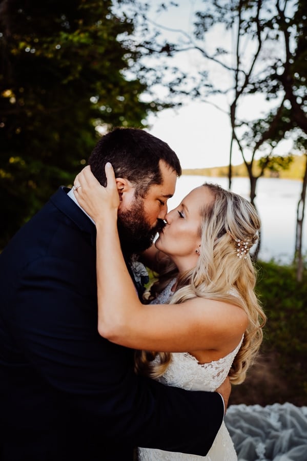 Bride with hands on grooms neck kissing in front of water and trees