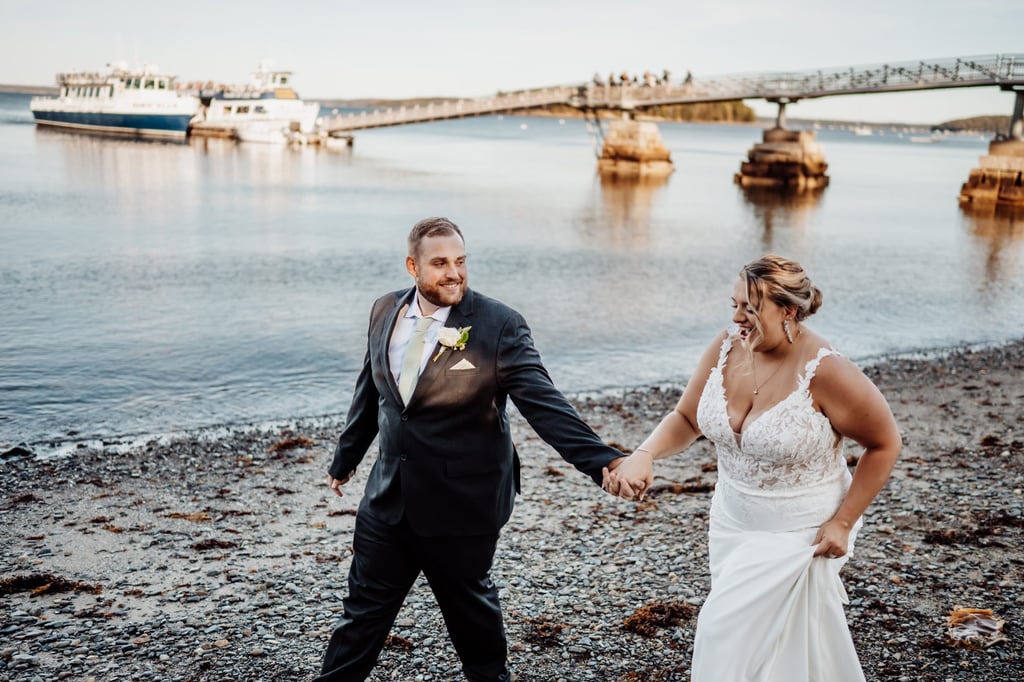 Bride and groom laughing and holding hands along the Bar Harbor shoreline with boats and the harbor bridge in the background during golden hour.