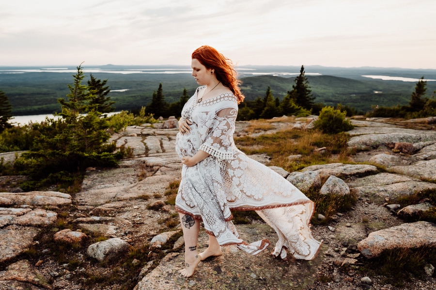 Pregnant woman holding belly in white dress on cadillac mountain