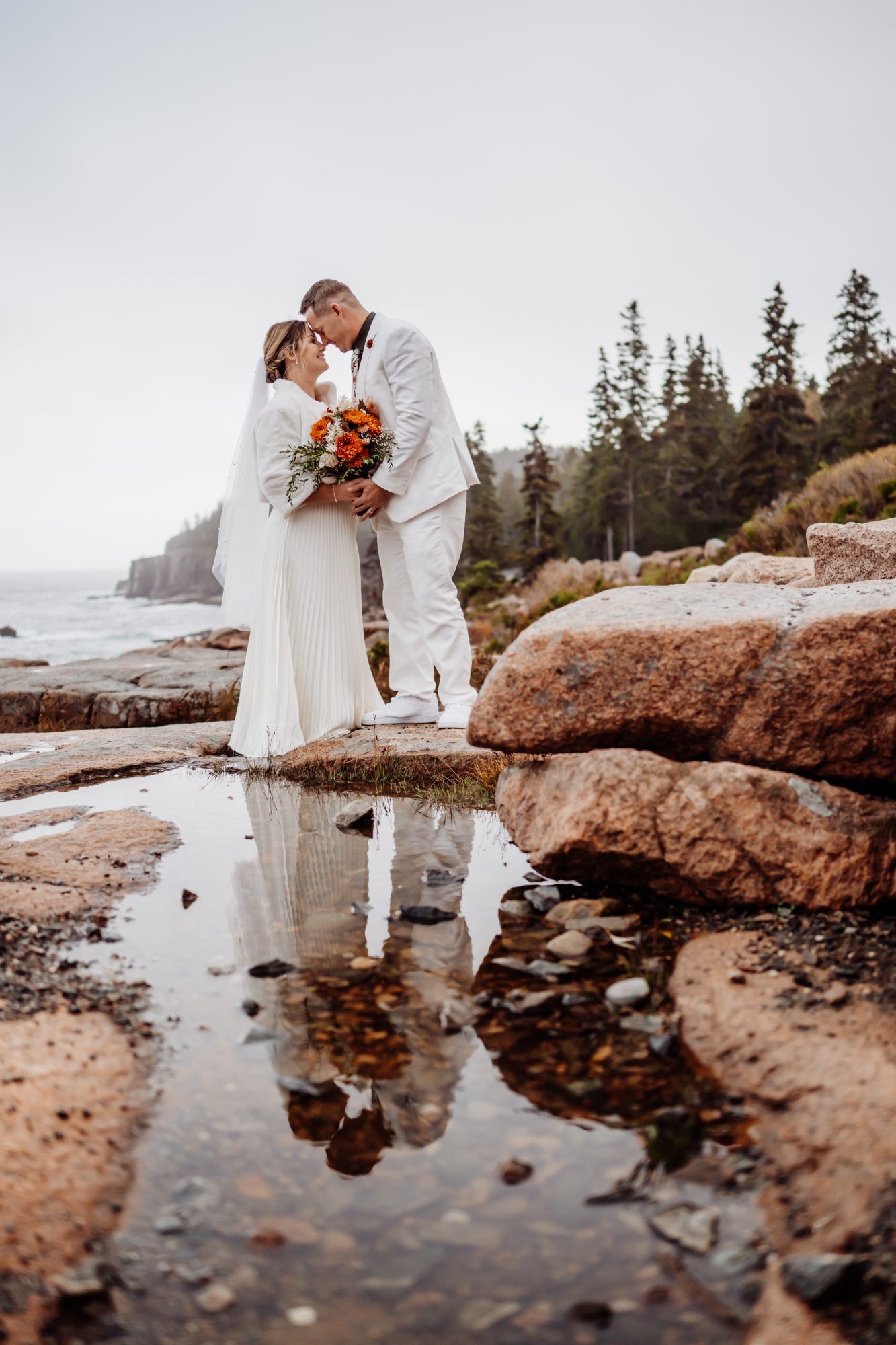 Bride and groom standing on coastal rocks with bouquet in Acadia National Park.