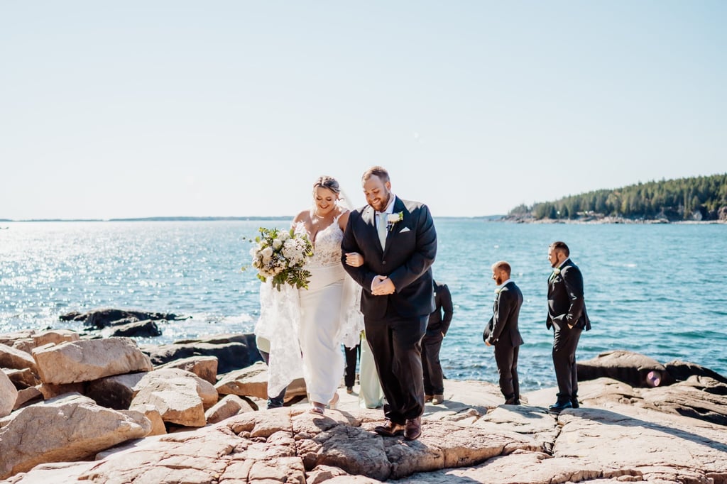 Newly married couple walking arm-in-arm along the rocky cliffs after their intimate Acadia National Park ceremony, smiling as guests and bridal party celebrate behind them.