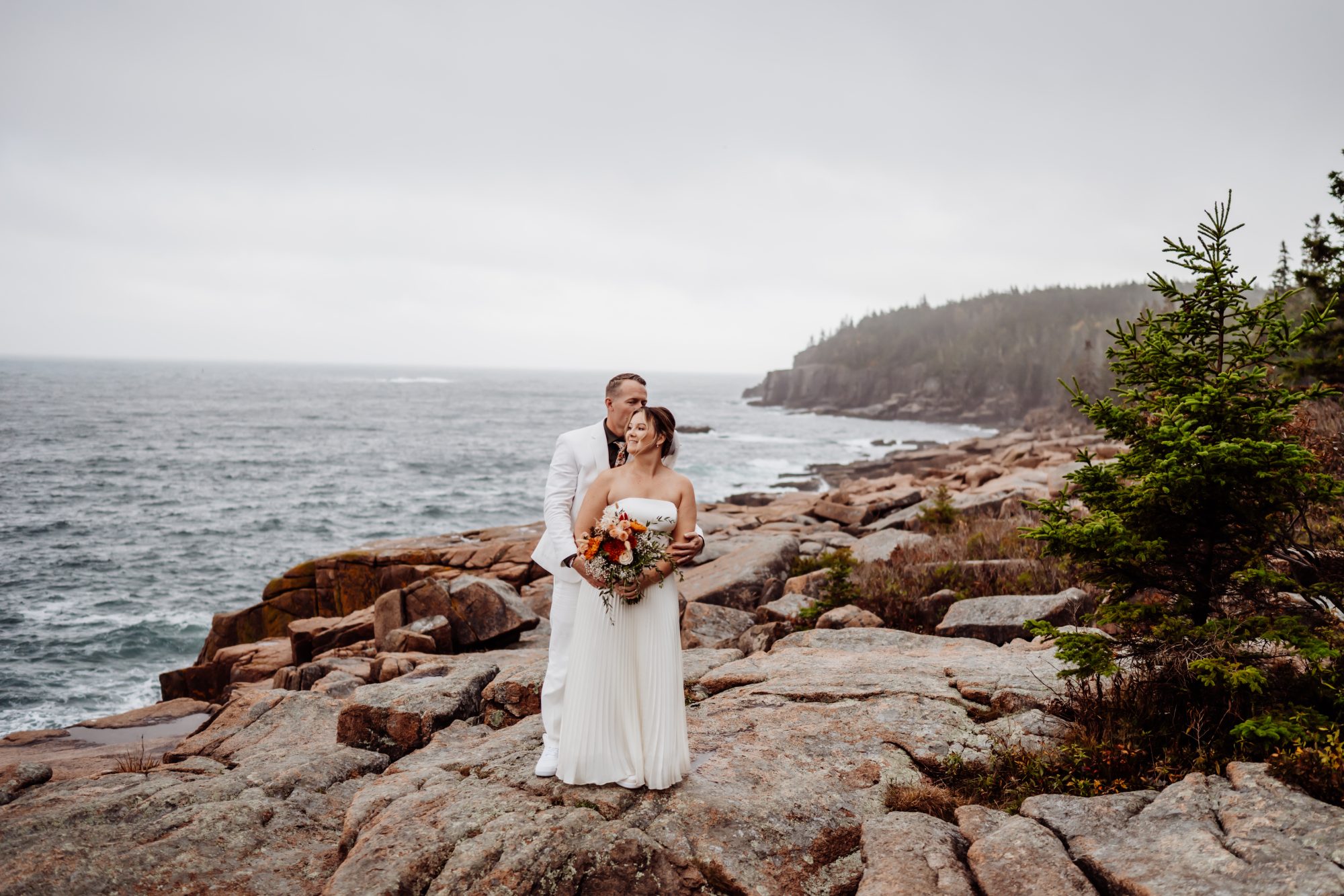 Newly married couple standing on rocky shoreline at Thunder Hole in Acadia National Park overlooking the Atlantic Ocean.
