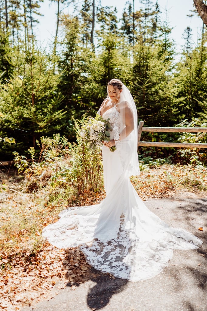 Bride standing on a wooded path in Acadia National Park, wearing a lace wedding gown with a long train and holding a lush white and green bouquet, photographed by Breezy Photography in Bar Harbor, Maine.