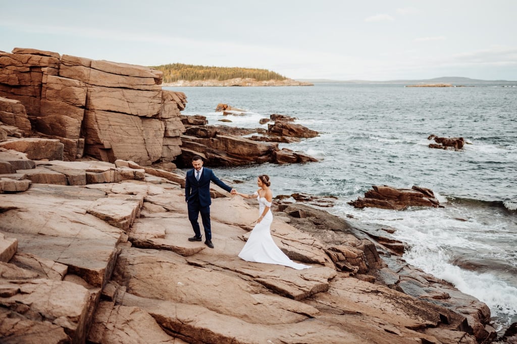 Bride and groom walking hand in hand along the granite cliffs at Schooner Head Overlook in Acadia National Park above the ocean.