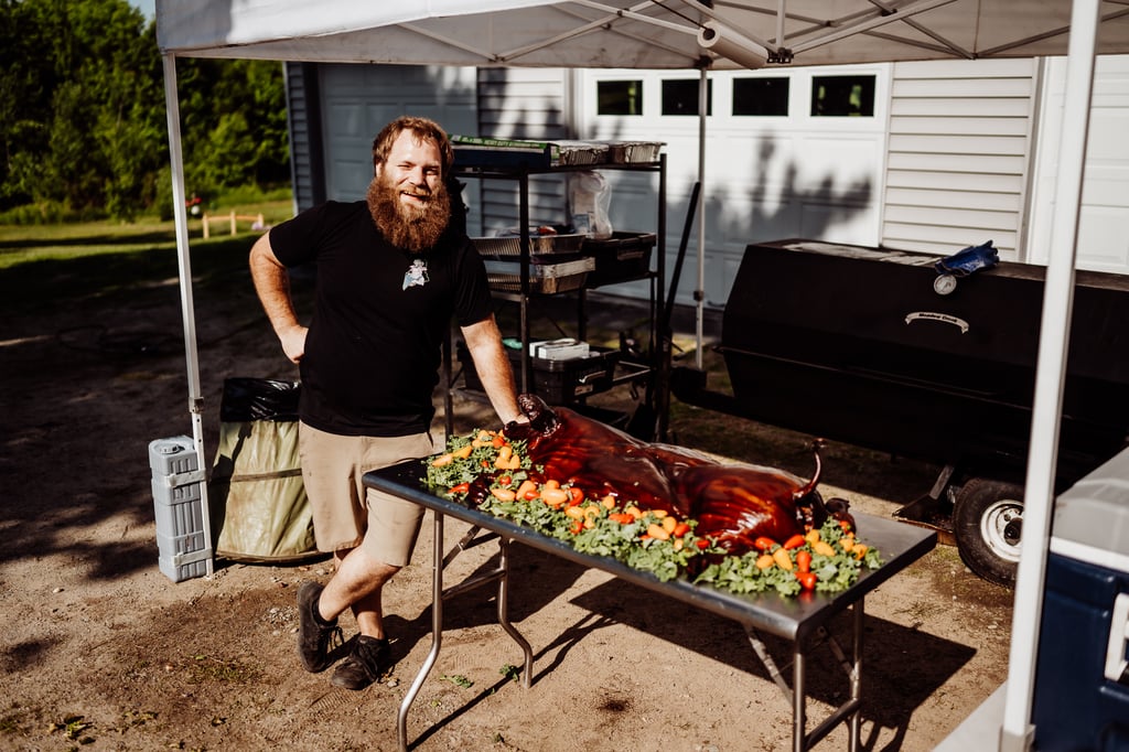 A man posing proudly next to a table displaying a roasted pig, ready for a wedding feast.