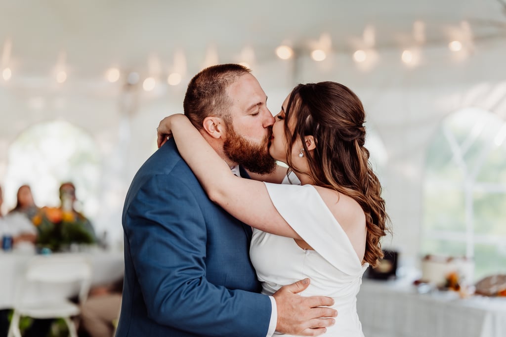 A newlywed couple shares a kiss during their wedding reception, with the groom in a blue suit and the bride in a white dress, under a tent decorated with soft string lights.