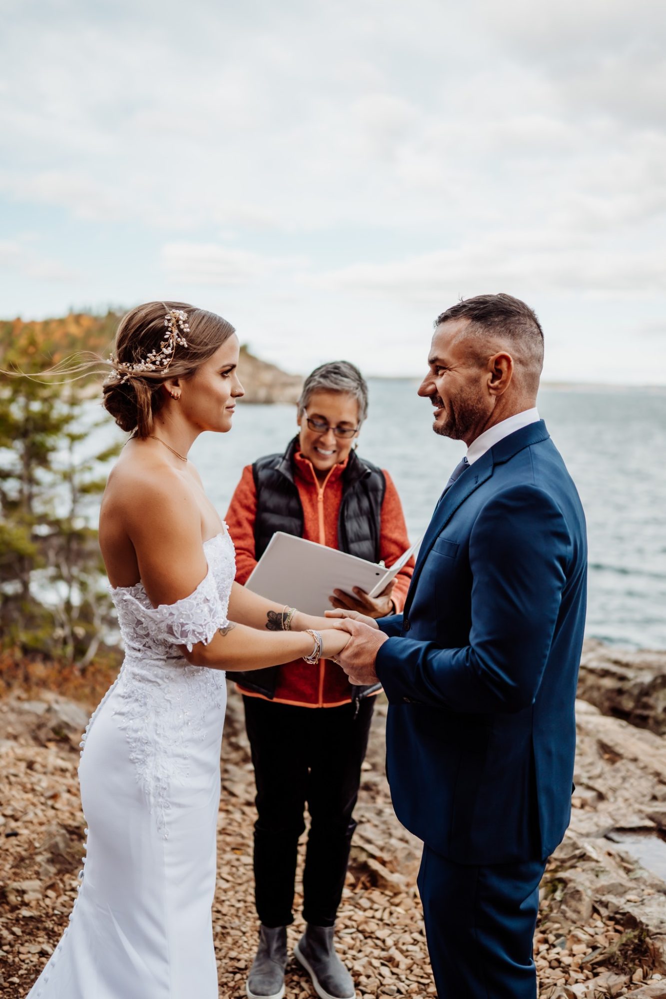 Couple smiling during their cliffside elopement ceremony at Schooner Head Overlook in Acadia National Park, officiant standing beside them with the Atlantic Ocean in the background.