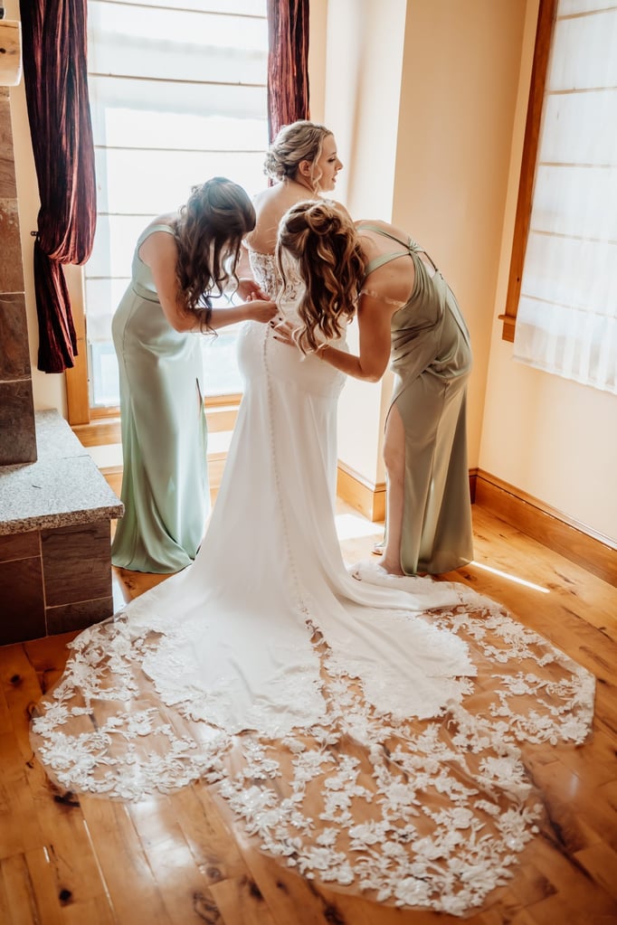 Bride in a lace-back gown being helped into her dress by two bridesmaids wearing sage green dresses, sunlight streaming through the window highlighting the long lace train.