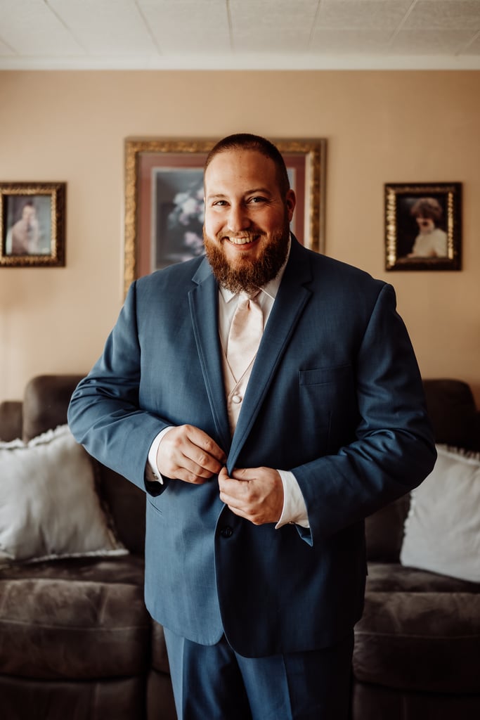 A groom in a blue suit smiles while buttoning his jacket, standing in a cozy, decorated living room