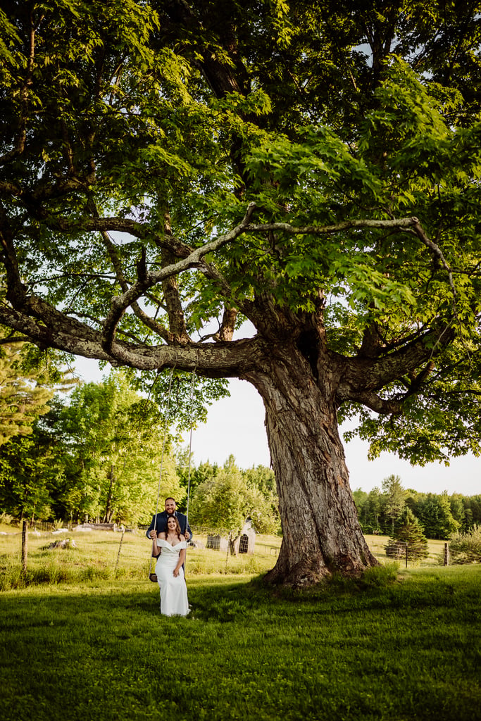 A couple stands together under a large, leafy tree in a lush, green meadow, with the man in a blue suit standing behind the woman in a white dress