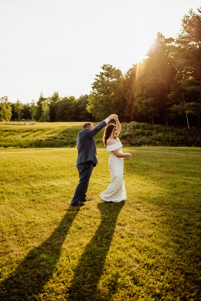 The groom twirls the bride in a field during sunset.