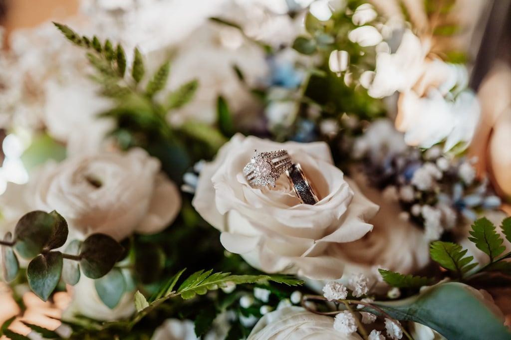 Close-up of wedding rings resting on a white rose within a lush bouquet of white flowers and greenery, softly lit with warm tones.