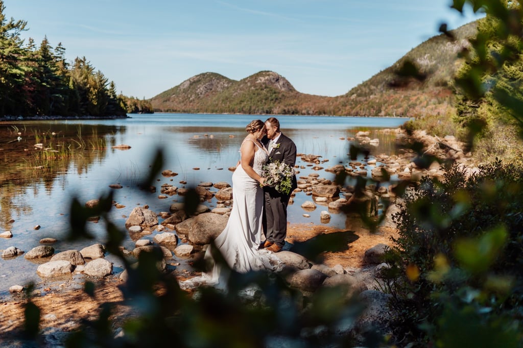 Bride and groom embracing by the water at Jordan Pond in Acadia National Park, framed by trees and mountain reflections.