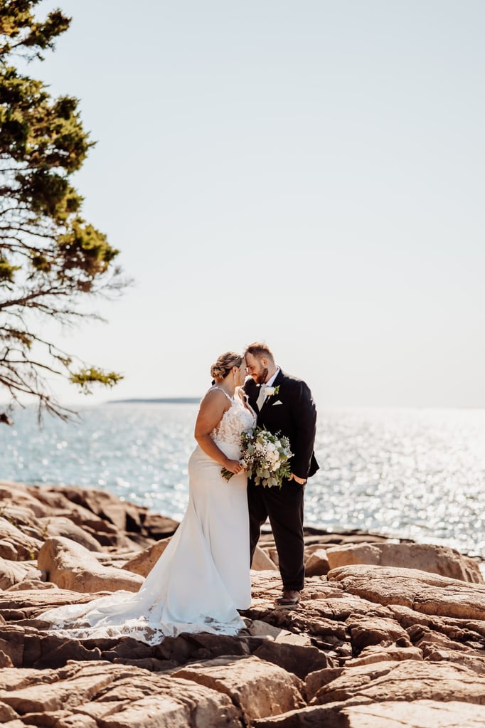 Bride and groom standing close together on the rugged coastline of Otter Point in Acadia National Park, sunlight sparkling on the water behind them.